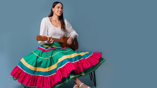 索尼娅·德·洛斯·桑托斯穿着一条彩色条纹大裙子坐在椅子上. 她拿着一个类似吉他的乐器，面带微笑.